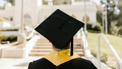 Back view of a graduate in a cap and gown standing outdoors facing a building.
